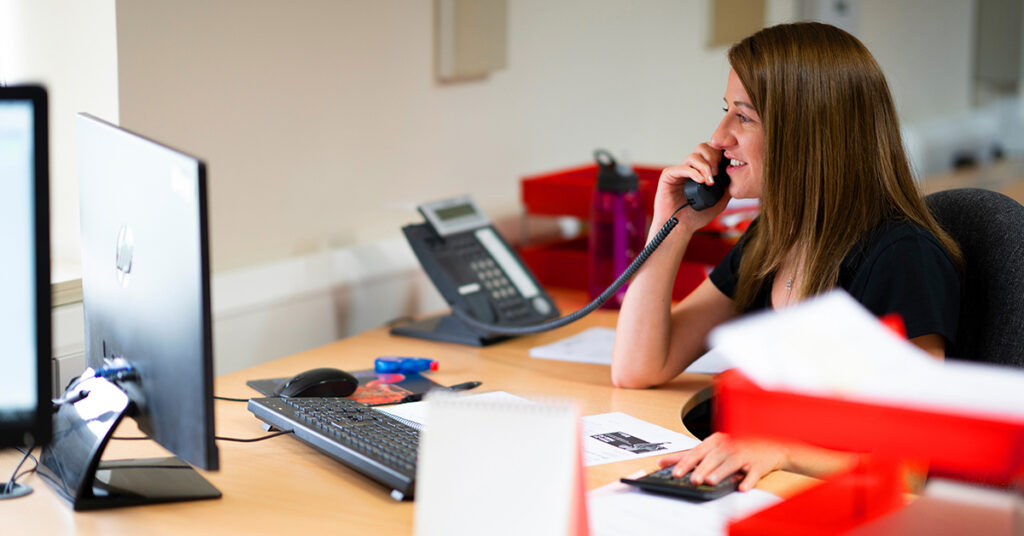 A lady sitting at a desk, smiling and on the phone - Red Rooster Lifting